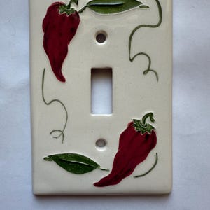 以下が含まれることがあります： クリーム色の背景に、2つの赤い唐辛子と緑の葉と茎、緑のつるのようなアクセントが描かれたセラミック製のスイッチカバー。 スイッチの開口部は中央にあります。
