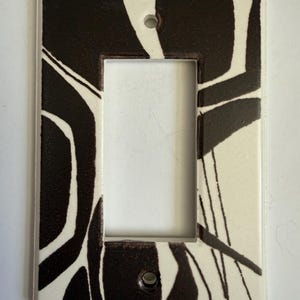 May include: Rectangular light switch plate with a black and white abstract design. The plate has a glossy finish and two screw holes. The design features black, curved lines and shapes against a white background.