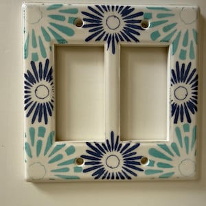 May include: A rectangular double light switch wall plate with a white base featuring a repeating hand-painted floral pattern in dark blue and light turquoise. The plate has two vertical rectangular openings and four screw holes for mounting.