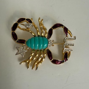 Op de afbeelding: Goudkleurige metalen Schorpioen broche met een turquoise middenstuk, gouden poten en een steraccent. De broche heeft een bordeauxrood emaille ontwerp en is versierd met heldere strass-steentjes.