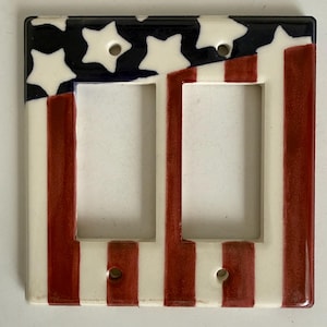 May include: A ceramic double light switch cover featuring a patriotic design. The top section has a blue background with white stars, while the lower section has red and white stripes. The cover has two openings for the switches.