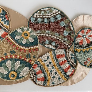 May include: A beaded Easter egg table runner with a beige satin backing. The eggs are in various sizes and colors, including teal, red, yellow, and brown, with intricate beadwork patterns. The runner is designed for decorative use.
