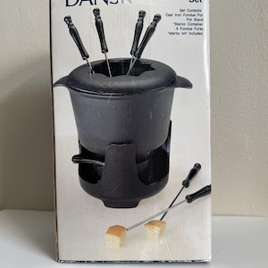 May include: A Dansk fondue set in a box featuring a black cast iron pot with a matching stand and six forks with black handles. The box displays the text Dansk Fondue Set, Set Contents: Cast Iron Fondue Pot, Pot Stand, Sterno Container, 6 Fondue Forks.