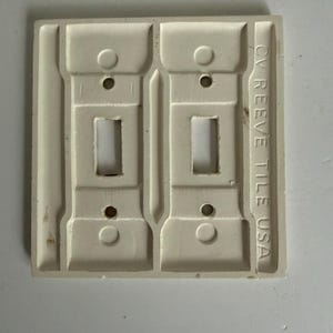 May include: A rectangular off-white double toggle light switch plate with a raised architectural design. The plate features two vertical cutouts for switches and four screw holes. The text CV REEVE TILE USA is embossed vertically on the right side.