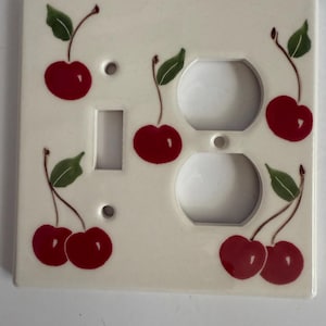 以下が含まれることがあります： クリーム色のセラミック製ライトスイッチカバー。さくらんぼのデザイン。トグルスイッチの開口部、2つのコンセント開口部があり、手描きの赤いさくらんぼと緑の葉で飾られています。家庭用装飾品。