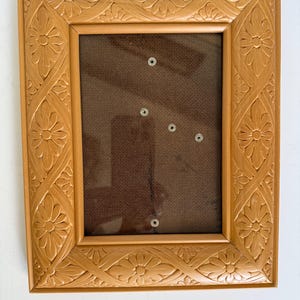 Może przedstawiać: Prostokątna, jasnobrązowa ramka na zdjęcia z kwiatowym wzorem. Rama ma wytłoczony wzór kwiatów i winorośli. Wnętrze ma ciemnobrązowe tło z kilkoma małymi, srebrnymi elementami mocującymi.