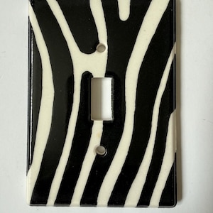May include: A decorative light switch cover with a black and white zebra stripe pattern. The cover has a rectangular shape with a single toggle switch opening and two screw holes.