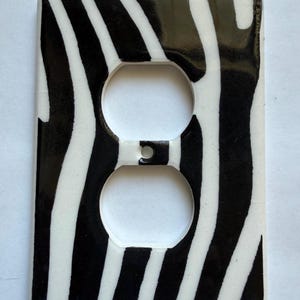 May include: A decorative electrical outlet cover featuring a zebra stripe design in black and white. The cover has two circular openings for the outlets and a small rectangular opening at the top.