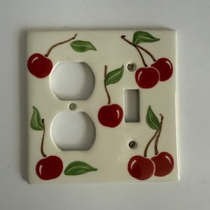 以下が含まれることがあります： クリーム色のセラミック製ライトスイッチカバー。手描きのチェリーのデザイン。カバーには2つのコンセント穴と1つのスイッチ開口部があります。赤いサクランボと緑の葉が表面に散らばっています。