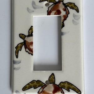 May include: A rectangular, off-white light switch cover featuring hand-painted sea turtles. The turtles have brown and green shells with black outlines. Gray wave-like shapes are painted around the turtles. The cover has a single rectangular opening.