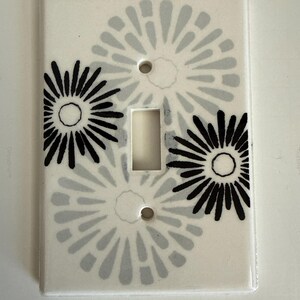 May include: A rectangular white light switch plate featuring a decorative pattern of three stylized flower-like bursts. Two of the floral designs are rendered in solid black, while the third design is depicted in a soft, muted light gray color.