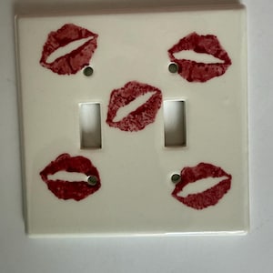 May include: A white double light switch cover with five red lipstick kiss prints. The cover has two rectangular switch openings and two screw holes. The lipstick prints are in a repeating pattern.