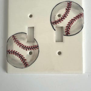 May include: A cream-colored, ceramic double light switch cover featuring two baseball designs. Each baseball is painted with gray shading and red stitching. The cover has two switch openings and two screw holes.