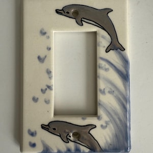 May include: A ceramic light switch cover featuring two gray dolphins leaping above blue waves. The cover has a rectangular shape with a central opening and is painted on a cream-colored background.