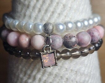 May include: A stack of three bracelets with a silver charm. The top bracelet is made of white pearls, the middle bracelet is made of pink and gray stones, and the bottom bracelet is made of clear stones.