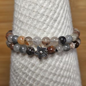 May include: Two beaded bracelets with a variety of earth-toned beads, including clear, brown, and gray. A silver-colored flower charm sits between the bracelets. The bracelets are displayed on a white, textured surface.