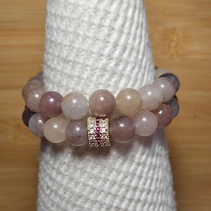 May include: Two beaded bracelets with light purple and tan-colored beads. The bracelets are held together by a gold-colored spacer with small red and clear stones. The bracelets are displayed on a white, textured surface.