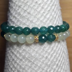 May include: Two beaded bracelets. The top bracelet features faceted, dark teal beads. The bottom bracelet has light green beads. Both bracelets have gold-colored spacers with small red accents. The bracelets are displayed on a white, textured surface.