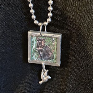 May include: A silver chain necklace with a rectangular pendant featuring a photo of a black cat in the grass. A small silver hummingbird charm hangs from the bottom of the pendant.