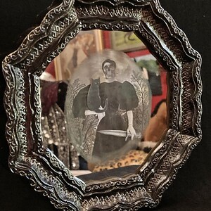 May include: A black and white portrait of a woman holding a black cat. The woman is wearing a long black dress and has a skull-like face. The portrait is framed in a black ornate frame.