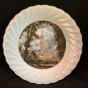 May include: A white ceramic plate with a gold rim features a printed image of a cathedral in a landscape setting. The image is surrounded by a white border.