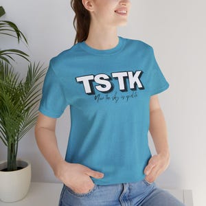 Pode incluir: Uma t-shirt de manga curta azul brilhante com o texto branco em negrito TSTK com uma sombra preta. Abaixo do texto principal, a frase Now the sky is opalite está impressa numa fonte cursiva preta mais pequena na zona do peito.
