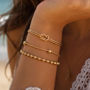 Peut inclure: Un ensemble de trois bracelets dorés portés au poignet. Le bracelet supérieur présente un nœud central, le bracelet du milieu est une fine chaîne avec deux petites perles sphériques, et le bracelet inférieur est composé d'une rangée de perles dorées.