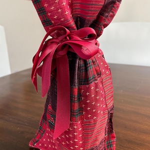 May include: A red fabric gift bag with a patchwork design. The bag features a variety of patterns, including stripes, plaid, and cross stitch, in shades of red and green. A matching red ribbon is tied in a bow at the top.