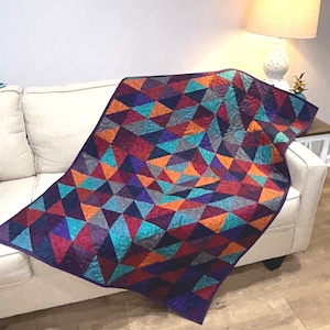 May include: A colorful quilt with a geometric pattern of triangles in shades of purple, teal, orange, and gray. The quilt is draped over a white sofa, showcasing its vibrant design and craftsmanship. The quilt's dimensions are not visible.