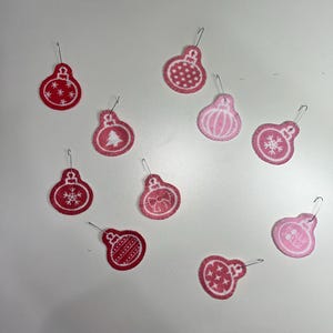 May include: A collection of red and pink Christmas ornament decorations with various designs, including snowflakes, a Christmas tree, and a bow. Each ornament has a silver hook for hanging. The ornaments are made of felt.