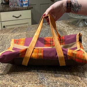 May include: A rectangular fabric carrier featuring a patchwork design of orange and yellow plaid squares alternating with solid plum-colored panels. The bag includes two sturdy mustard-yellow fabric handles and a solid dark brown base panel.