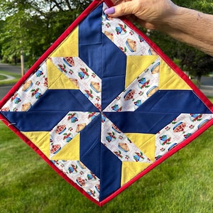 May include: A square quilted hot pad with a red border. The design features a starburst pattern with navy blue, yellow, and pie-themed fabric. The hot pad is held up against a green background.
