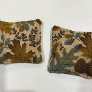 May include: Two square fabric coasters with a fall leaf pattern in shades of brown, green, and blue on a beige background. The coasters are edged with black stitching. The design includes oak leaves, pinecones, and other foliage.
