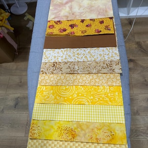 May include: A collection of yellow and gold fabric swatches arranged in a vertical stack. The fabrics display various patterns, including floral, geometric, and abstract designs. The color palette ranges from pale yellow to deep gold, creating a visually appealing display of textures and hues.