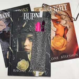 May include: A book with a gray and black cover featuring a woman's face and a white rose. A gray fabric bookmark with a black swirl pattern holds two pens. A pair of glasses rests on the book.