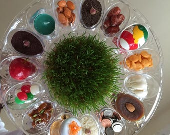 May include: A clear plastic tray with 12 compartments, each containing a different item. The items include: wheatgrass, almonds, dried fruit, coins, a painted egg, a small jar of honey, a small jar of sugar, a small jar of salt, a small jar of rice, a small jar of lentils, a small jar of dried beans, and a small jar of dried herbs.