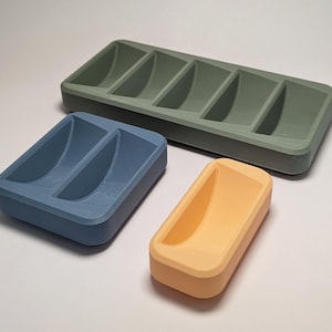 Puede incluir: Tres bandejas impresas en 3D de diferentes tamaños y colores. La más grande es una bandeja rectangular verde oliva con cinco compartimentos curvos. Una bandeja azul más pequeña tiene dos compartimentos, y una melocotón tiene uno.