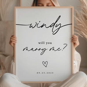 May include: A framed white sign with the words "windy will you marry me?" in black script. A heart and the date 09.25.2023 are below the text. The sign is held by a person.