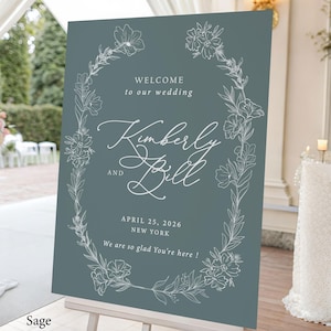 May include: A sage green rectangular wedding welcome sign featuring a white line-art floral wreath border. The text reads: WELCOME to our wedding, Kimberly AND Bill, APRIL 25, 2026, NEW YORK, We are so glad You're here ! The sign is on a white stand.