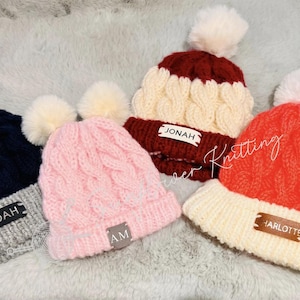 May include: Four cable-knit beanies with white pom-poms. Colors include navy blue with gray brim, pink, burgundy and cream, and orange with cream brim. Each beanie has a tag with a name or initials: NOAH, AM, JONAH, and HARLOTTE.