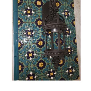 Op de afbeelding: Een donkerbruine smeedijzeren wandlamp met een lantaarndesign is gemonteerd op een decoratieve tegelwand. De tegels hebben een herhalend patroon van blauwe, gele en witte bloemmotieven.