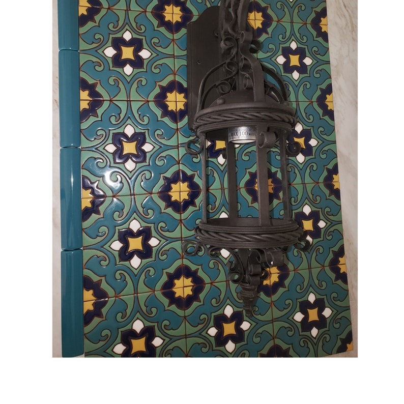 Op de afbeelding: Een donkerbruine smeedijzeren wandlamp met een lantaarndesign is gemonteerd op een decoratieve tegelwand. De tegels hebben een herhalend patroon van blauwe, gele en witte bloemmotieven.