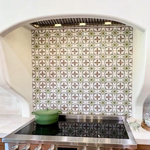 May include: A kitchen scene featuring a stainless steel range with a black cooktop and a green pot. The backsplash is tiled with a repeating pattern of green, red, and white crosses and floral designs. The range hood is above the cooktop.
