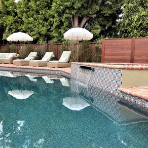 Op de afbeelding: Een serene buitenruimte met een turquoise zwembad dat witte parasols weerspiegelt. Rieten ligstoelen met groene kussens staan bij het zwembad. Decoratieve tegels omzomen het zwembad en een houten hek zorgt voor privacy.