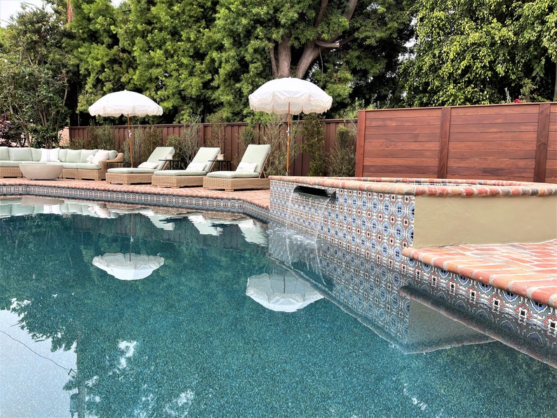 Op de afbeelding: Een serene buitenruimte met een turquoise zwembad dat witte parasols weerspiegelt. Rieten ligstoelen met groene kussens staan bij het zwembad. Decoratieve tegels omzomen het zwembad en een houten hek zorgt voor privacy.
