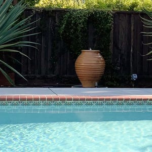 Op de afbeelding: Een grote, bruine, geribbelde keramische pot staat op een betonnen richel bij een zwembad. Het zwembad heeft turquoise water en een decoratieve tegelrand. Palmachtige planten en een houten hek staan op de achtergrond.