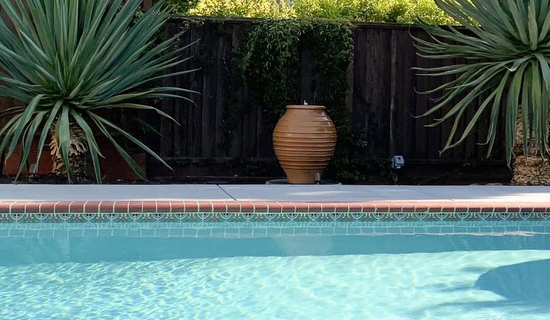 Op de afbeelding: Een grote, bruine, geribbelde keramische pot staat op een betonnen richel bij een zwembad. Het zwembad heeft turquoise water en een decoratieve tegelrand. Palmachtige planten en een houten hek staan op de achtergrond.