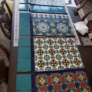 May include: An assortment of decorative ceramic tiles in various patterns and colors. The tiles feature floral and geometric designs in shades of blue, teal, yellow, and red. Some tiles are solid teal, while others have intricate patterns.