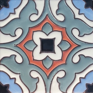 Op de afbeelding: Vierkante keramische tegel met een symmetrisch bloemmotief. De tegel heeft een centraal zwart vierkant omgeven door een witte, vierbladige bloemvorm. Het ontwerp bevat teal, oranje en blauwe elementen, omlijnd in zwart, tegen een witte achtergrond.