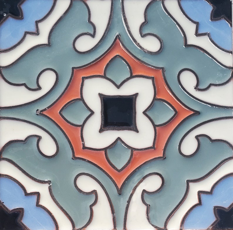 Op de afbeelding: Vierkante keramische tegel met een symmetrisch bloemmotief. De tegel heeft een centraal zwart vierkant omgeven door een witte, vierbladige bloemvorm. Het ontwerp bevat teal, oranje en blauwe elementen, omlijnd in zwart, tegen een witte achtergrond.